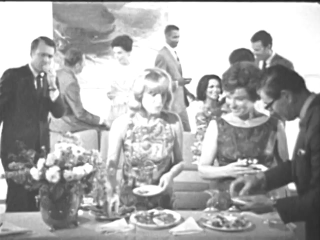 The still from the 'Ad Council PSA - Presidents Council on Youth Opportunity' features a social gathering where several people are interacting in a well-decorated setting. The foreground shows two women engaging with food on a table, while others, dressed in formal attire, mingle in the background. The atmosphere appears lively, embodying a vintage social event typical of the 1960s. There are floral arrangements and plates of food present, suggesting a focus on community and social connection.