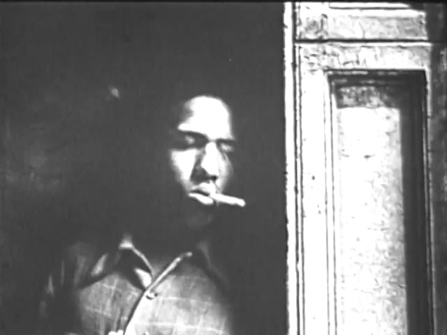 The still features a man standing in a doorway, partially illuminated by light. He is depicted with a cigarette in his mouth, giving a contemplative or relaxed expression. The image is in black and white, creating a stark contrast and a gritty urban feel, which aligns with the themes often explored in public service announcements aimed at youth. The overall mood suggests a moment of reflection or pause in a potentially challenging environment.