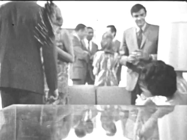 The still from the 'Ad Council PSA - Presidents Council on Youth Opportunity' features a group of individuals in a casual gathering. In the foreground, a woman in a seated position is seen with her head down, while others engage in conversation in the background. They are dressed in formal attire, suggesting a professional or formal setting, likely indicating a meeting focused on youth opportunities. The overall ambiance is social, with a focus on interaction among the attendees.