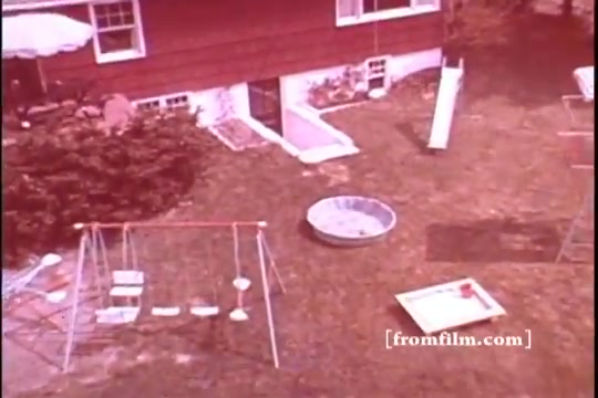 The still depicts a backyard scene from the 1970s, featuring a red house with large windows. In the foreground, a swing set with several swings can be seen, along with a small sandbox and a kiddie pool. A slide is positioned nearby, suggesting a family-friendly environment. The overall color tone has a vintage look, characteristic of 16mm prints from that era.