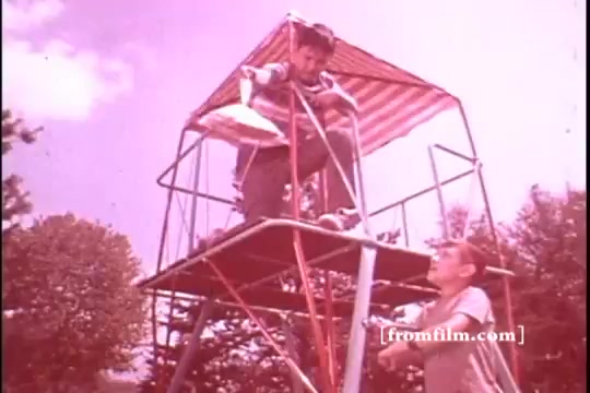 The still features two boys outdoors, set against a bright sky. One boy is perched atop a playground structure, holding what appears to be a bag of Baggies Food Storage Bags, while the other stands below, looking up at him. The scene captures a playful moment of childhood curiosity, framed in a soft, pink hue typical of 1970s film stock. The overall atmosphere is lighthearted and nostalgic, emphasizing the theme of outdoor play and the utility of Baggies.