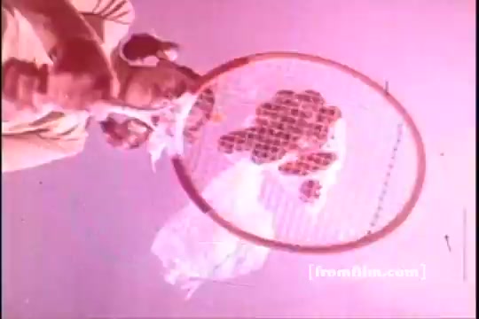 The 16mm still from the 1970s Baggies Food Storage Bags advertisement features a playful scene where an individual, likely a child, holds a tennis racket beneath a floating bag filled with food. The image has a vibrant, pinkish hue, adding a whimsical touch to the lighthearted promotion of the storage bags. The focus is on the interaction between the racket and the bag, emphasizing the product's versatility and fun.