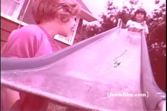 The still features two children on a playground slide, typical of the 1970s era. One child, with blonde hair, is positioned at the bottom of the slide wearing a pink shirt, while another child, dressed in white, stands at the top. The background includes a house and trees, lending a nostalgic, summery vibe. The color tone is warm, reflecting the film style of that time. The scene captures a moment of playful anticipation, embodying the carefree spirit of childhood in the 1970s.