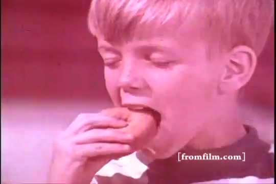 The still captures a young boy, likely from the 1970s, about to take a bite from a cookie or small pastry. He has short blonde hair and is wearing a striped shirt. The color palette appears warm, with a pinkish hue typical of older film prints. The image is focused on the boy's joyful expression, emphasizing the theme of food enjoyment, possibly showcasing the convenience and appeal of Baggies Food Storage Bags.