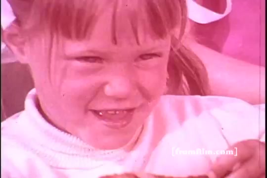 The still features a young girl with a playful smile, likely enjoying a snack. She has light brown hair styled with bangs and is wearing a white sweater. The image has a nostalgic, slightly faded quality typical of 1970s film, emphasizing warm tones. In the background, other figures are present, suggesting a casual, joyful setting, possibly related to family or friends. The overall mood conveys a sense of fun and comfort associated with food storage and sharing.