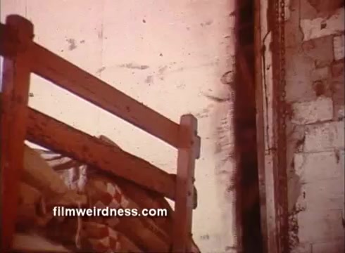 The still depicts a simple interior scene, likely a room with a wooden bed frame and a loosely made bed featuring patterned bedding. The walls appear weathered and worn, hinting at a rustic or old-fashioned setting. Subtle color tones and film grain suggest it is from a vintage film, contributing to a nostalgic atmosphere reflective of the era.