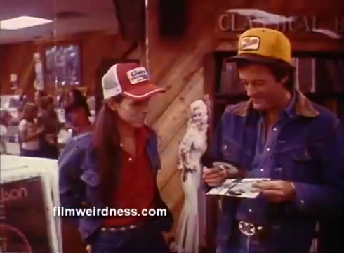 The still from the 'Outlaw Blues' trailer features two characters engaging in conversation in a record store. One character wears a red shirt and denim jacket, while the other dons a yellow cap and a blue shirt. The store's interior has wooden paneling and various items on display, including records and a visible figure in the background. The scene embodies a casual, nostalgic vibe typical of the film's era.