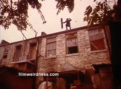 The still features a gritty urban setting, showcasing a dilapidated building with a stone facade. A figure can be seen on the roof, balancing precariously with a wooden plank, suggesting themes of danger or rebellion. The overall color tone is warm, hinting at a nostalgic or vintage style, characteristic of the era in which 'Outlaw Blues' was made.