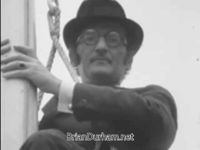 The still from 'A Matter of Life and Breath' (1968) features a flagpole sitter who expresses a fear of germs. He is dressed formally, wearing a suit and a hat, and appears to be perched on top of a flagpole. The scene captures a moment of quirky humor and societal commentary, typical of the film's exploration of odd characters and their unique fears.