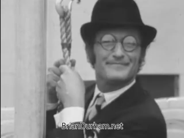 The still from 'A Matter of Life and Breath' features a flagpole sitter who appears cheerful despite his fear of germs. He is dressed in a suit and bowler hat, holding onto a rope attached to the flagpole. The scene captures the juxtaposition of his smile and the underlying anxiety about cleanliness, likely reflecting the film's themes surrounding life's challenges and personal fears. The black-and-white imagery emphasizes the era and adds to the comedic tone.