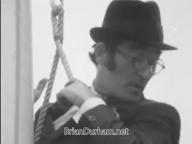 The still from 'A Matter of Life and Breath' (1968) features a flagpole sitter who appears anxious and is concerned about germs. He is wearing a bowler hat and glasses, and sits precariously on the flagpole, looking down with a worried expression. The image captures a moment of comedic tension, reflecting themes of anxiety and societal pressures prevalent in the film.
