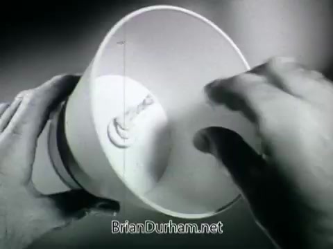 The still from the 'Plantivater commercial - 'A Max Klein Product'' features a close-up view of a person's hands interacting with a planter, specifically a Plantivater. The planter appears to be designed with a unique interior structure, likely aimed at promoting the health and growth of houseplants.  The transcript includes a voiceover introducing the product as a home for beautiful house plants, while a woman expresses her lack of confidence in caring for plants. The voiceover reassures her that the Plantivater can help, suggesting a user-friendly experience. The black and white aesthetic emphasizes the retro feel of the advertisement, showcasing both the product's design and the messaging aimed at plant enthusiasts or those hesitant about their gardening skills.