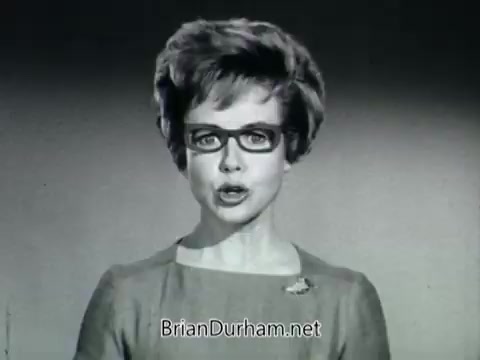 The still from the 'Plantivater commercial' features a woman speaking directly to the camera, portraying confidence and authority. She has a classic 1960s look, with styled hair and glasses, dressed in a simple, elegant outfit. The background is a neutral tone, accentuating her presence.  In the context of the commercial, she addresses concerns about plant care, emphasizing that the Plantivater is suitable even for those who consider themselves not good with plants. The voiceover complements her dialogue, enhancing the message about the ease of using Plantivaters for beautiful houseplants. The overall tone feels persuasive, aimed at reassuring potential customers about their ability to care for plants using this product.