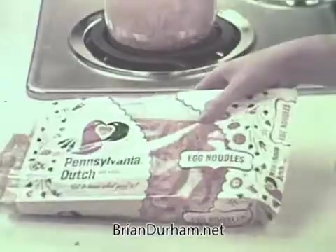 The still features a hand holding a package of Pennsylvania Dutch Egg Noodles, prominently displayed in the foreground. In the background, a pot appears to be boiling on the stove. The packaging is colorful, with a distinctive design highlighting the brand name. The setting suggests a home kitchen environment typical of the 1960s, emphasizing a domestic, family-oriented theme.