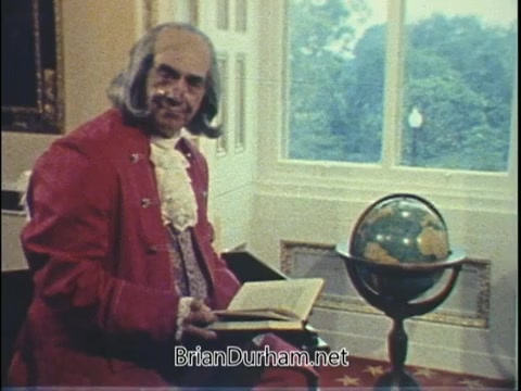 The still features a man dressed in 18th-century attire, complete with a wig and a bright red coat, seated next to a globe on a stand. He holds an open book, appearing engaged in a presentation. The background suggests a well-lit room with large windows, hinting at a historical or educational context. The overall aesthetic reflects a vintage style typical of government PSAs during that era.