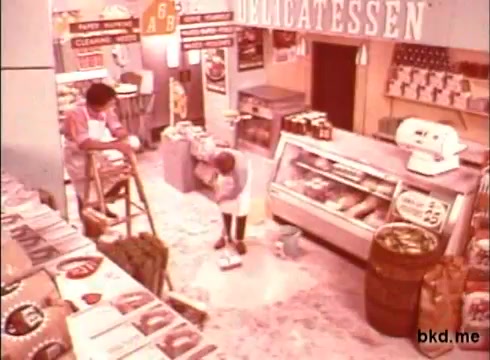 The still depicts a bustling delicatessen in the 1970s, featuring a worker on a ladder taking notes or doing inventory. Another person is on the floor, possibly cleaning, emphasizing a focus on cleanliness. The shop is filled with various products displayed on shelves and in a glass case, highlighting the vibrant, colorful atmosphere typical of that era. The overall scene conveys a sense of hard work in a professional food setting.
