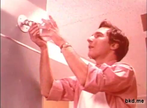 The still from the 1970s 'Spic and Span' commercial features a man in a collared shirt, seemingly in the process of cleaning or preparing a surface. He appears focused and engaged, holding a cleaning product or sponge. The environment suggests a domestic setting, likely a kitchen or bathroom, characterized by a simple, utilitarian design typical of that era. The color palette is warm, reflecting the 70s aesthetic.
