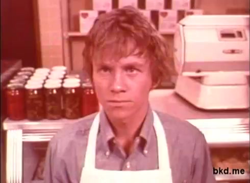 The still features a young man with tousled hair, wearing a light blue shirt and an apron. He has a serious expression and is positioned in a store or diner setting. Behind him, there are cans lined up on a shelf and a vintage cash register. The overall vibe reflects the style and aesthetics of 1970s advertisements.