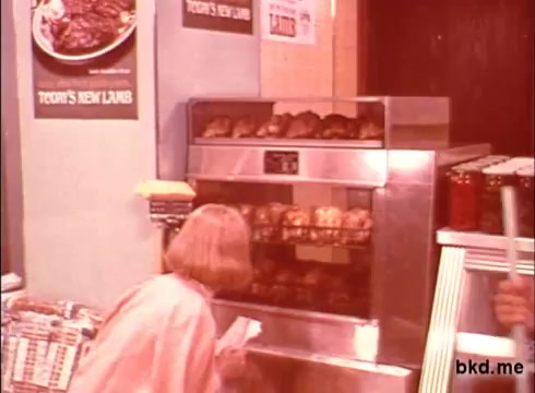 The still captures a scene from a 1970s commercial for Spic and Span. It features a woman in a light-colored outfit, possibly interacting with a display of roasted meats in a glass-fronted oven. The background includes posters advertising 'Today's New Lambs,' suggesting a focus on fresh food. The overall aesthetic reflects the vibrant colors and style typical of that era, with an emphasis on food preparation and presentation.