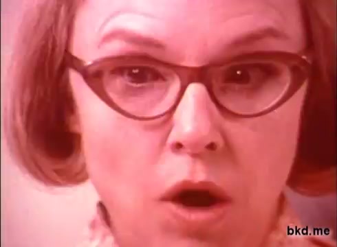 The still features a woman with a surprised expression, possibly reacting to something impressive. She's wearing large, round glasses and has short, light-colored hair. The colors are vivid, typical of 1970s commercial aesthetics, and the close-up emphasizes her facial features and emotions. The background is blurred, drawing attention to her reaction.