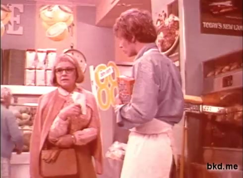 The still features a scene from a 1970s Spic and Span commercial. It depicts a woman with glasses and a coat, holding a bag, engaged in conversation with a man in an apron, who is holding a container. The background features various grocery items, giving a domestic or store setting. The color tone exhibits a soft, vintage quality typical of 16mm prints from that era.