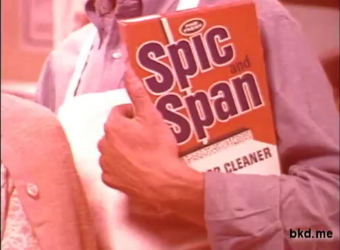 The still features a person dressed in casual attire, holding a box of Spic and Span, a well-known cleaning product from the 1970s. The box is prominently displayed, highlighting its vibrant orange color and branding. The setting has a warm, nostalgic tone typical of advertising from that era. The individual appears engaged, suggesting a positive association with the product.