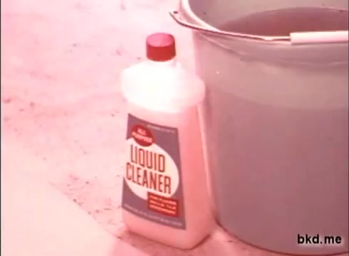 The still features a vintage 16mm commercial for Spic and Span from the 1970s. In the foreground, there is a bottle of Spic and Span Liquid Cleaner displaying its label prominently. A bucket of water is positioned nearby, emphasizing the cleaning theme. The colors are typical of the era, with a slightly faded look indicative of older film prints. The setting appears to be a simple indoor space, likely intended to showcase the product's usage in cleaning.