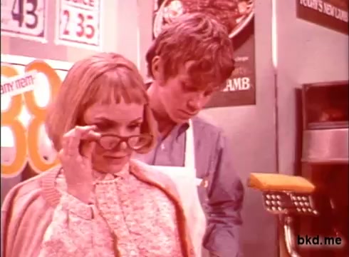 The still from the 'Spic and Span' commercial features a woman with short blonde hair and glasses, adjusting her glasses as she looks at a young man in an apron. The young man appears to be engaged with something on a counter, likely at a food-related setting. The overall color tone is characteristic of 1970s film, displaying warm hues and a retro vibe. Background elements suggest a commercial kitchen or market environment.