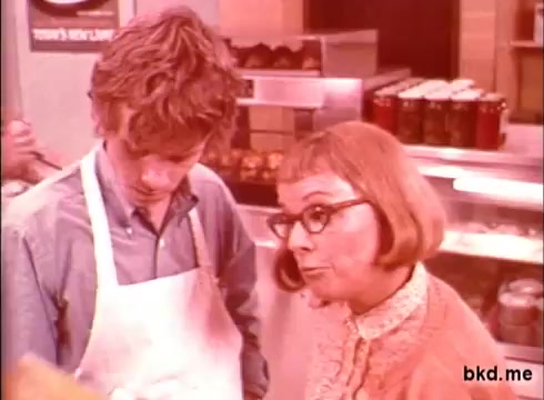 The still features two characters in a casual setting, likely a food establishment. One character, a young man in an apron, appears to be engaged in conversation with an older woman wearing glasses and a cardigan. The warm, soft color palette suggests a nostalgic feel typical of 1970s commercials. The background shows various food items, emphasizing a theme of cleanliness and care in food preparation.