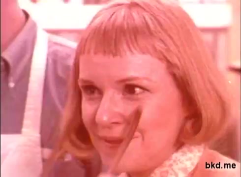 The still features a woman with a short, styled haircut and a warm expression, likely in a kitchen setting. She holds a brush, possibly for applying a cleaning product, which aligns with the commercial's promotional theme. The color tone is warm and soft, characteristic of 1970s advertising aesthetics. A person is partially visible in the background, suggesting a collaborative context.
