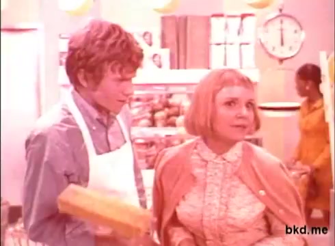 The still from the 'Spic and Span' commercial features two characters in a brightly lit store setting. One character, a young man in a white apron, holds a yellow box, while the other, a woman with a short blonde hairstyle, appears to be engaging with him, possibly in conversation or persuasion. The color tone has a warm, pink hue, characteristic of 1970s aesthetics, and the background includes shelves and a clock, suggesting a grocery store environment. Another figure can be seen in the background, adding to the lively atmosphere.