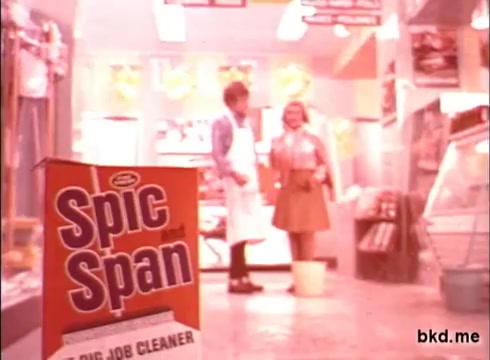 The still features a bright, colorful commercial for Spic and Span from the 1970s. In the foreground, a box of Spic and Span cleaner is prominently displayed. In the background, two people, a man in an apron and a woman, appear to be engaged in conversation, likely in a retail setting. The overall aesthetic captures the vibrant and visually engaging style typical of advertising from that era.