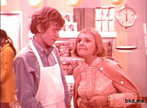 The still from the 'Spic and Span' commercial features a scene set in a brightly colored grocery store. A young man in an apron appears engaged in conversation with a woman who has a bob hairstyle and is wearing a light-colored cardigan. The overall aesthetic is vibrant and slightly retro, typical of 1970s advertising, conveying a playful interaction amid a backdrop of grocery items.