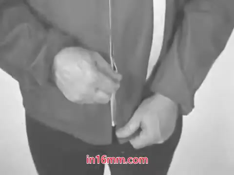 The 16mm still from the advertisement features a man demonstrating the action of zipping up a jacket. The scene is likely in a simple, uncluttered setting, focusing on the man's hands as he pulls the zipper up. This visual pairs with the voiceover announcing 'Total,' an international gasoline brand. The ad emphasizes the product's global experience and innovation in gasoline blending.