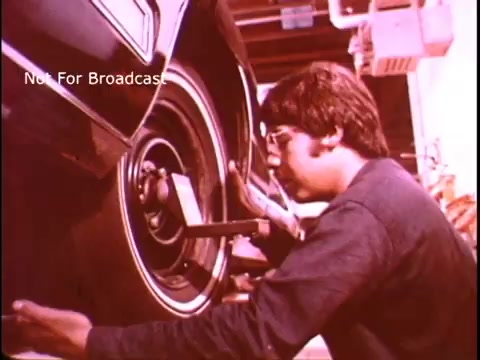 The still depicts a young man working on a car tire, using a tool to adjust or inspect it. He appears focused on his task, showcasing hands-on skills. The setting suggests a vocational or automotive training environment, typical of Job Corps PSAs from the 1970s, which aimed to promote skill development and job readiness. The color tone has a warm, vintage quality typical of that era.