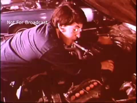 The still from the '70s Job Corps PSA features a young person working intently under the hood of a car. Dressed in a blue work shirt, they are focused on the engine, suggesting hands-on vocational training. The scene emphasizes skill development and the importance of job readiness in automotive fields. The background suggests an auto shop environment, typical of the era's vocational education themes.