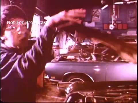 The still features a person gesturing energetically, possibly explaining a task or demonstrating a technique in a workshop setting. In the background, a blue car is partially visible amidst various tools and equipment, suggesting an automotive repair context. The overall atmosphere reflects a hands-on approach to vocational training, likely emphasizing the value of the Job Corps program in the 1970s.