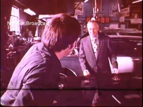 The still shows a scene from a 1970s Job Corps public service announcement. A young man, dressed casually, is facing an older man in formal attire who appears to be engaging him in conversation. The setting is a workshop or garage, with tools and cars visible in the background, conveying a vocational training environment. The overall tone suggests mentorship or guidance in a professional setting.