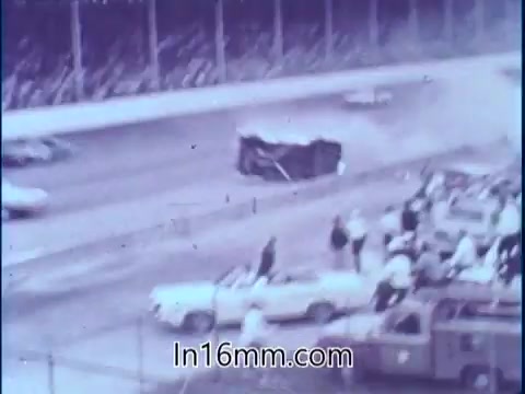 The still captures a dramatic moment from a NASCAR race, showing a car that has flipped over on the track during a chaotic scene at Darlington Raceway. Spectators in the stands and on the sidelines appear alarmed as debris is visible in the aftermath of the crash. This image illustrates the unpredictability and danger of stock car racing, highlighting the importance of safety equipment, as emphasized by driver Sam McQuaig.