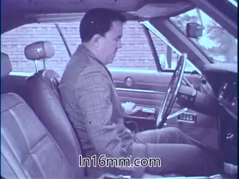 The still shows Sam McQuaig sitting in the driver's seat of a car. He appears focused, likely preparing to drive. The interior is classic, featuring a simple dashboard and front seat design. The scene suggests a moment of contemplation before action, aligning with the theme of automotive safety and the importance of being prepared for unexpected events, as highlighted in his PSA.