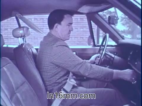 The still depicts a man, Sam McQuaig, seated in a car with a focused expression, likely preparing to drive. He is wearing a seatbelt, emphasizing the importance of safety equipment in vehicles. The car's interior suggests a classic model, and the background features a brick wall, indicating a casual environment. This visual reinforces the PSA's message about the significance of safety gear in preventing serious injuries during unexpected incidents, such as car crashes.