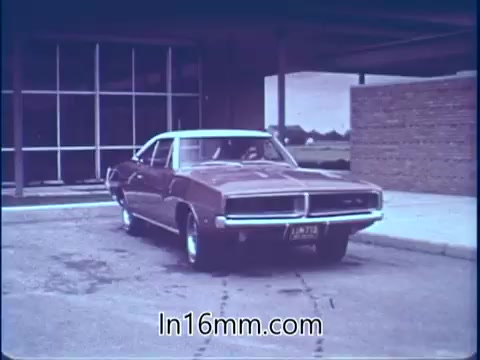 The still features a vintage brown car parked outside a building with large windows. The atmosphere appears calm and subdued. The context references Sam McQuaig, a stock car driver who emphasizes the importance of safety equipment after experiencing a crash during a race at Darlington Raceway. It highlights the unexpected nature of automotive incidents and the critical role of safety measures.