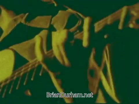 The still captures a close-up of a musician playing an instrument, possibly a guitar or similar string instrument. The image is tinted with a yellow and green hue, creating a vintage, artistic effect. This visual style reflects the time period and context of the 'Pittsburgh Symphony Promenades Commercial', emphasizing the performance aspect of the symphony. The focus on the hands and instrument enhances the dynamic and intimate feel of live music.