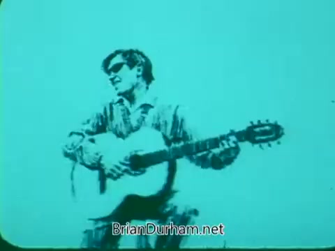 The still features a musician playing an acoustic guitar, set against a turquoise background. The figure is wearing sunglasses and appears to be engaged in a performance. The overall aesthetic suggests a stylized or retro look, typical of 16mm film. This image likely captures the vibrant and artistic spirit of the Pittsburgh Symphony Promenades.