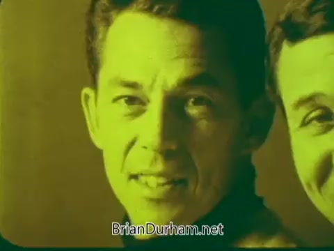 The still features two men smiling, highlighting their faces with a greenish tint. The shot captures a friendly, warm atmosphere, likely aimed at promoting the Pittsburgh Symphony Promenades. The focus is on their expressions, suggesting enthusiasm and engagement with the event.