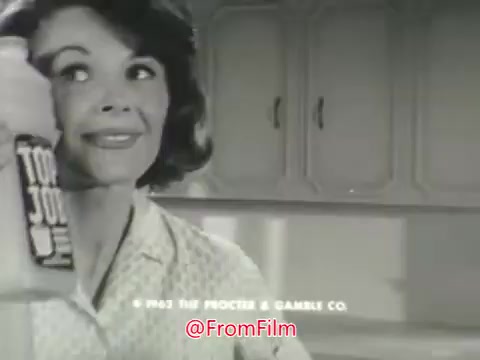 In this 16mm still from the 'Top Job cleaner with ammonia!' commercial, a woman holds a bottle of Top Job cleaner, smiling confidently. The background features a tidy kitchen, emphasizing cleanliness. The dialogue reflects a conversation with a man, discussing the appearance of floors, leading to a voice-over highlighting Top Job as a new cleaning product from Procter and Gamble. The tone is upbeat, showcasing the product's effectiveness in brightening dull floors.