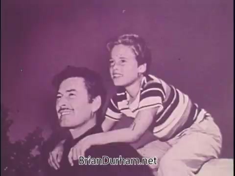 The still from the PSA features Jackie Cooper, likely seated at a location outdoors. He appears with a young boy, who is wearing a striped shirt. The boy is playfully seated on Cooper's shoulders, both looking cheerful. The background is softly blurred, giving a nostalgic feel to the image. The overall tone suggests a warm moment, capturing a sense of joy and innocence, which contrasts with the serious subject of Sean Flynn's disappearance. The image is part of a campaign to bring awareness to Flynn's fate, emphasizing the human side of this tragic narrative.