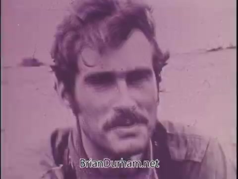 The still from the PSA features a close-up of a man with a mustache and tousled hair, appearing serious and engaged. The background is blurred, suggesting he is outdoors, possibly in a war-torn or rugged environment. The purple tint gives the image a vintage feel, emphasizing its historical context. The voiceover, delivered by Jackie Cooper, introduces the segment and focuses on the story of Sean Flynn, highlighting his role as a photojournalist and the circumstances surrounding his disappearance in Cambodia in 1970. The overall tone is reflective and somber, aiming to raise awareness about Flynn's legacy and the challenges faced by journalists in conflict zones.