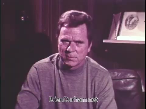 The still features Jackie Cooper in a close-up, seated against a backdrop that suggests a cozy indoor setting, possibly a study or living room. He wears a simple gray sweater, and his expression appears serious and reflective, likely conveying the gravity of the subject matter. There are books or artifacts visible on a shelf behind him, hinting at a thoughtful environment. The overall mood seems somber, as he addresses the audience about Sean Flynn's disappearance and the impact of such a loss. The visual style is characteristic of the period, adding to the historical context of the message.