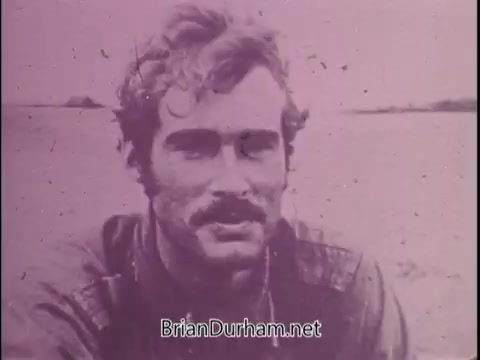 The still appears to capture a moment from a public service announcement featuring Jackie Cooper discussing Sean Flynn, the son of actor Errol Flynn, who was a photojournalist in Vietnam. The image likely portrays Sean Flynn with a serious expression, possibly reflecting his adventurous yet perilous life. The color tone suggests a vintage quality, emphasizing the era in which these events occurred. The voiceover by Jackie Cooper adds a somber context, highlighting Flynn's disappearance in Cambodia during the conflict and his eventual declaration of death.